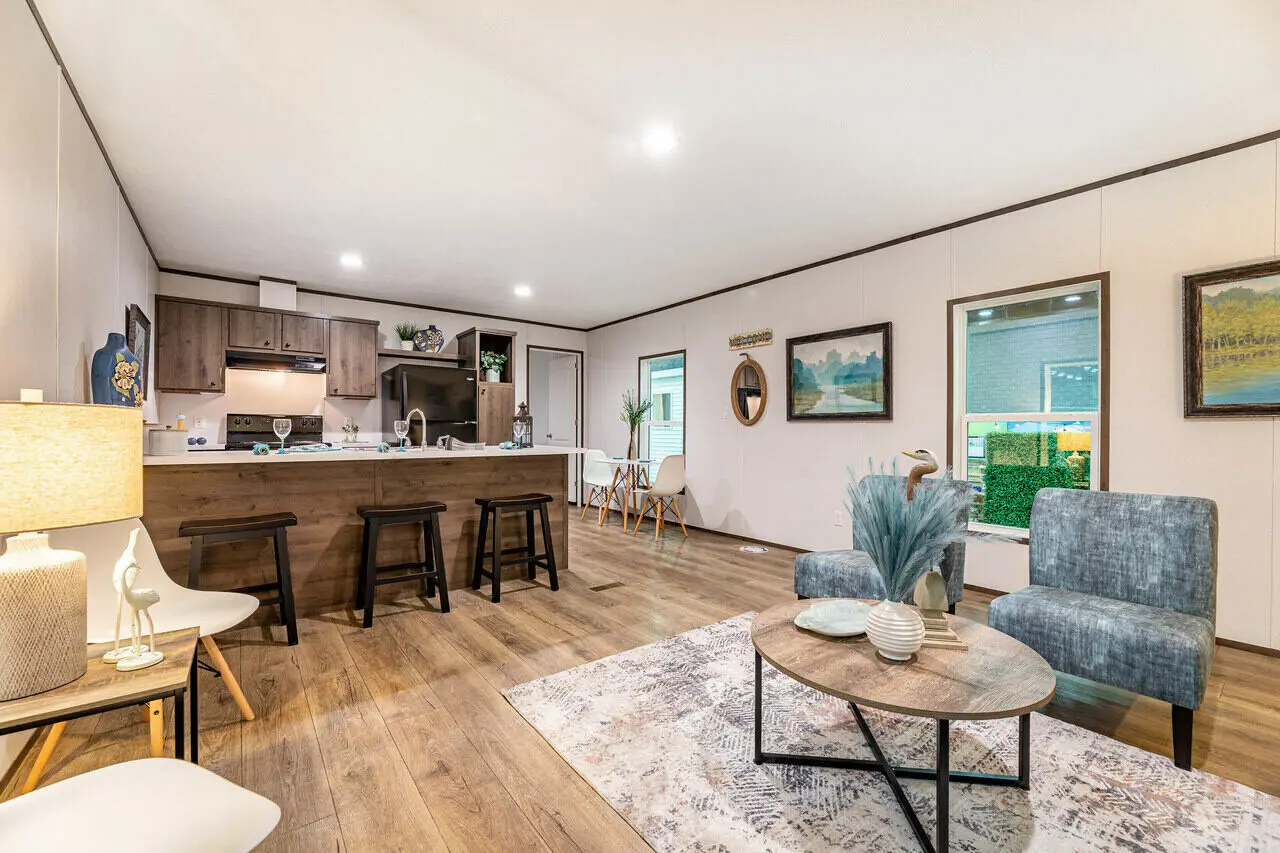 A modern living space with wooden flooring features a seating area with chairs and a coffee table in the foreground. The background displays a kitchen with a dining bar and stools, along with various decor items.