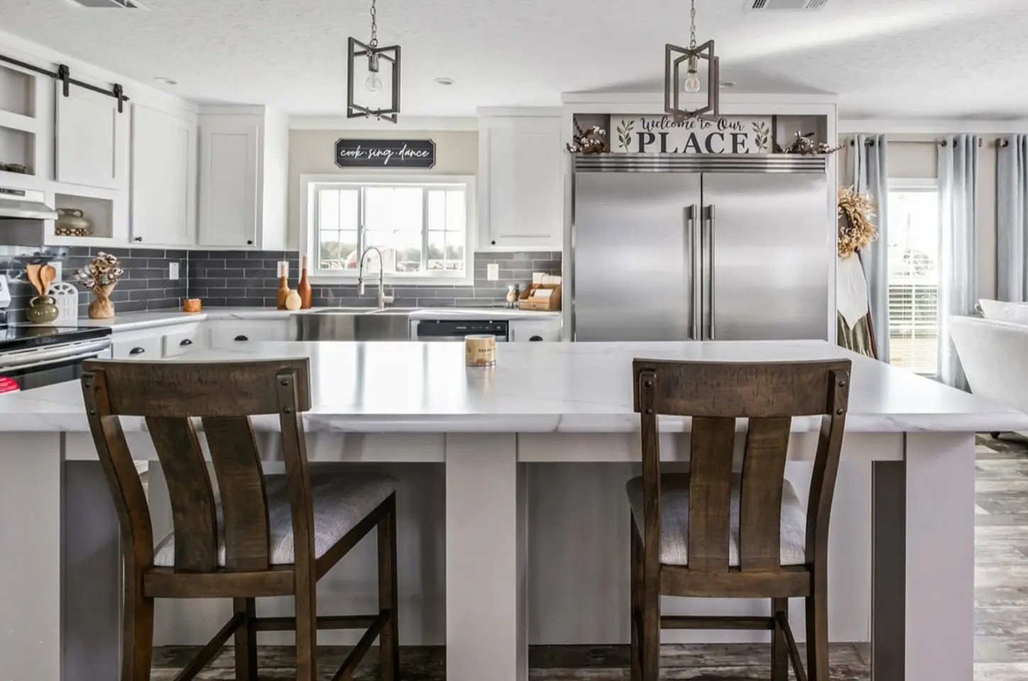 Modern kitchen interior with white counters, stainless steel appliances, wooden chairs, and decorative lighting.