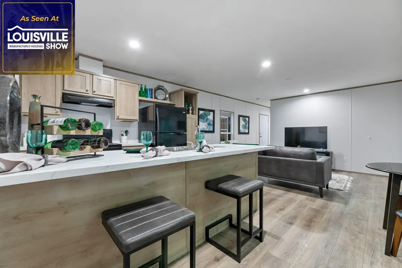 Modern kitchen and living area with a breakfast bar, two stools, light wood cabinets, and stainless steel appliances. The living area features a sofa, television, and wall art. "As Seen at Louisville" sign.