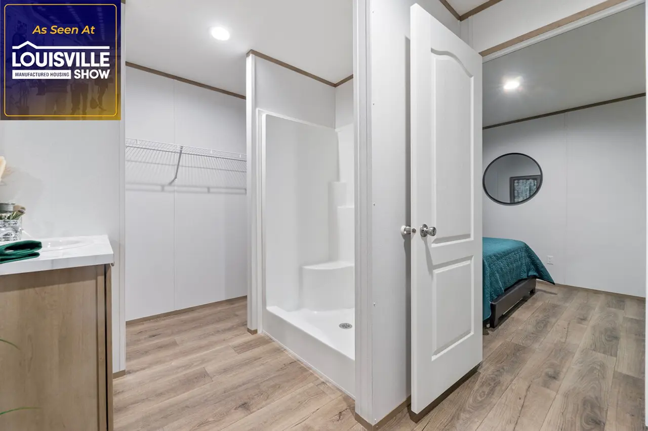 A small bathroom with an open shower stall next to a bedroom. A "Louisville Show" logo is in the upper left corner. The room features light walls, wooden floors, and an open closet with wire shelving.