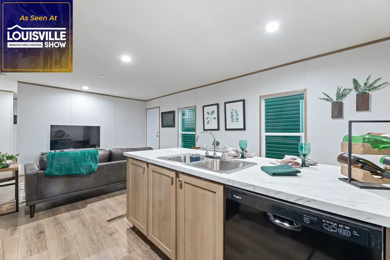 Modern living room and kitchen in a manufactured home with wooden cabinetry, marble countertop, green accents, sofa, TV, framed artwork on white walls, and green blinds on windows.