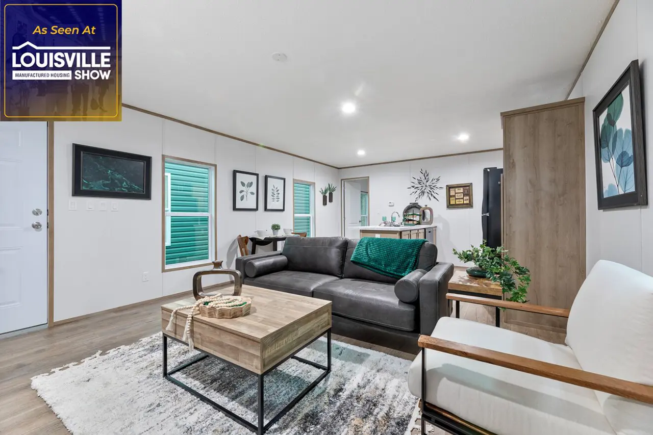 A modern living room features a gray sofa with a green throw, a wooden coffee table, framed art on the walls, and large windows with green blinds. A sign in the upper left reads, "As Seen At Louisville Show.