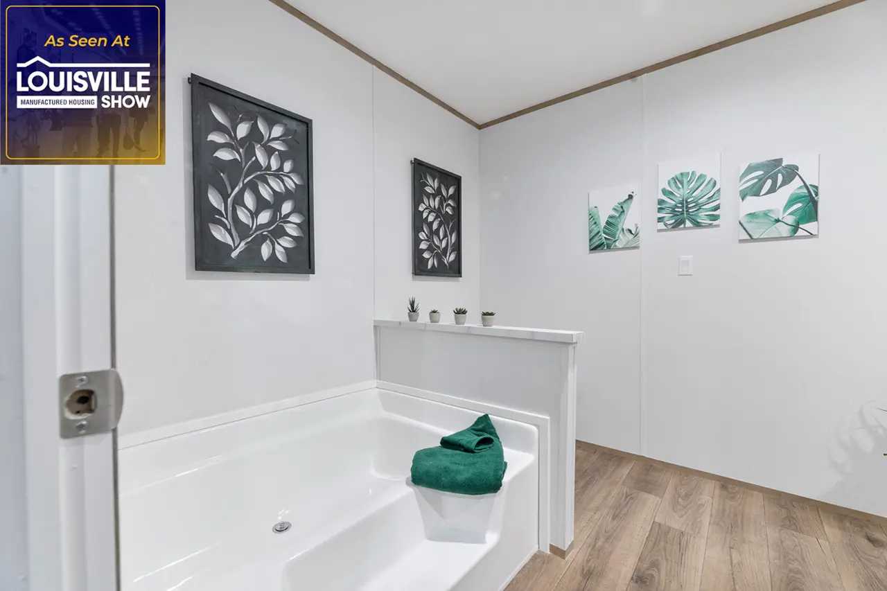 A clean, modern bathroom with a white tub, green towel, and leaf-themed artwork on the walls. The Louisville Manufactured Housing Show logo is displayed in the upper left corner.