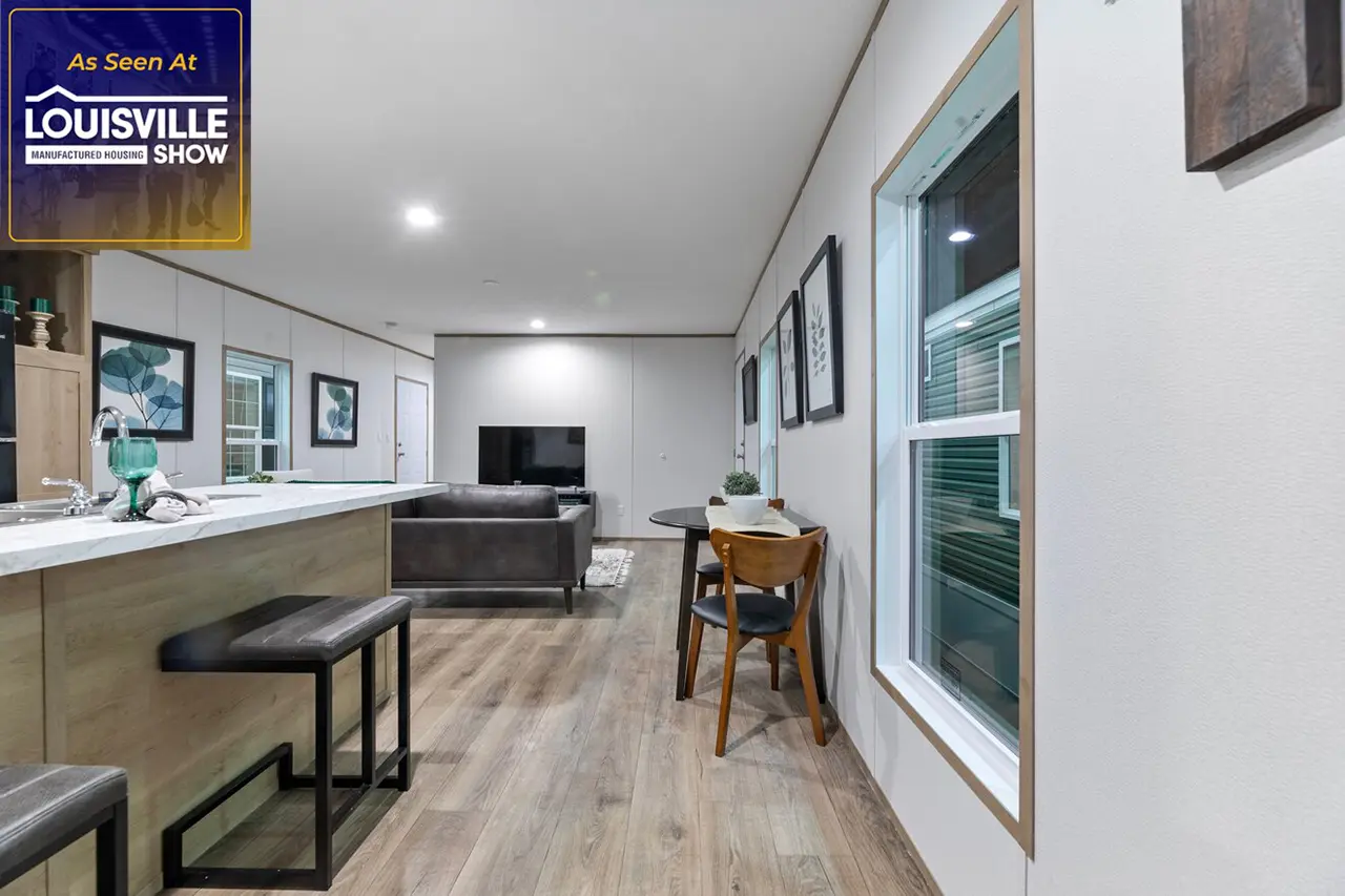 Modern living area with hardwood flooring, a kitchen island with barstools, a dining table with chairs, and a gray sofa facing a large TV. A window is visible on the right side.