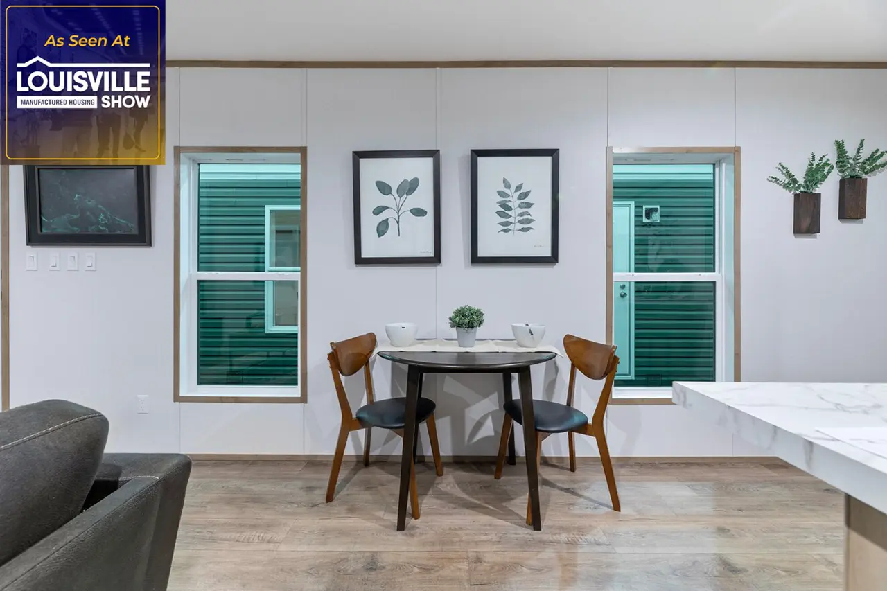 Small dining area with a round table, two chairs, and a potted plant. Two framed botanical prints and two windows with green shutters are in the background. Plaque reads "As Seen At Louisville Show.
