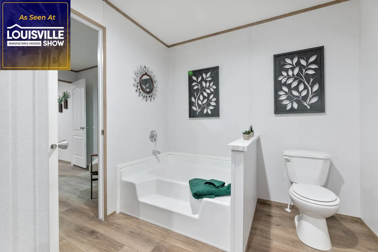 Modern bathroom with a white soaking tub, green towel, toilet, and decorative wall art. The bathroom has wooden flooring and connects to another room. A sign indicates it was featured at the Louisville Show.