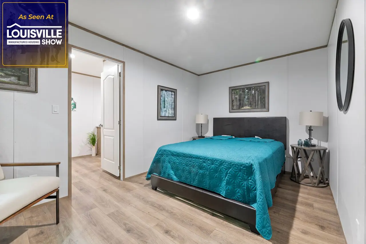 A minimalist bedroom with a double bed and blue bedspread, light wood flooring, and white walls. There's a lamp on a bedside table, framed pictures on the wall, and an open door leading to another room.