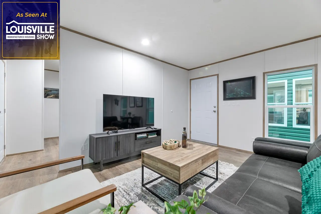 A modern living room features a wall-mounted TV, a coffee table, a couch, a chair, and wall decor. A sign reads "As Seen at Louisville Manufactured Housing Show.