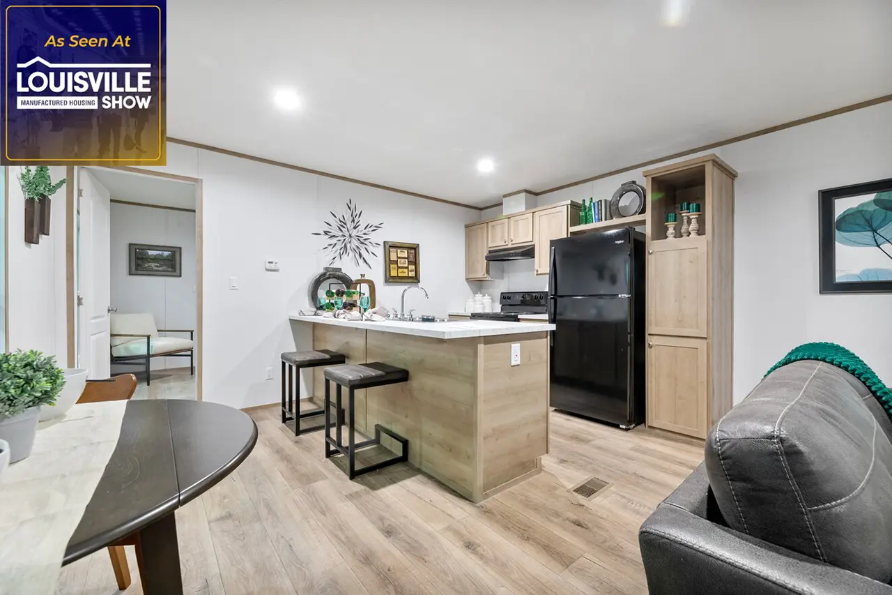 A modern kitchen with light wood cabinets, a black refrigerator, and a central island with two stools. There is a small dining area in the foreground and a living area visible to the right.