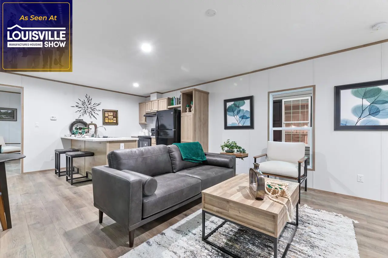 A modern living room with a gray sofa, an armchair, a wooden coffee table, and decorative items. The kitchen and dining area are in the background. A sign reads "As Seen At Louisville Manufactured Housing Show.