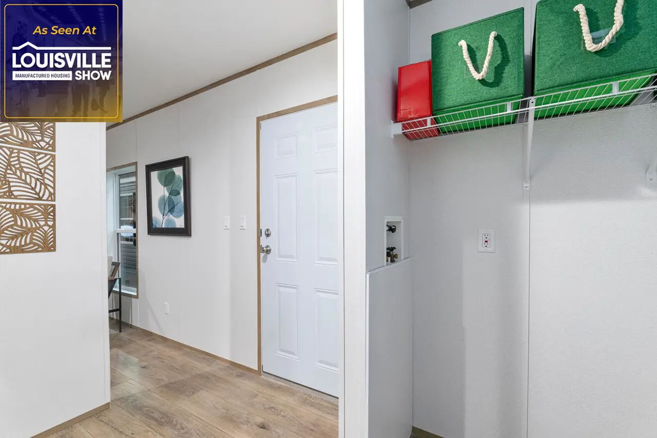 A hallway with wooden flooring leading to a white door, a wall-mounted shelf with green and red storage bins, and a framed picture on the wall. A sign for the "Louisville Show" is in the top left corner.