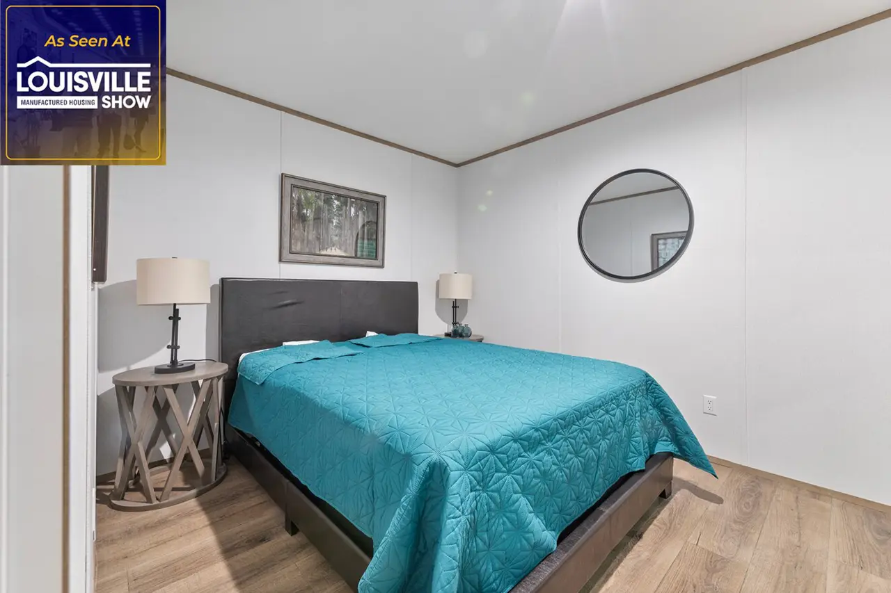 A bedroom with a double bed covered in a teal quilt, two bedside tables with lamps, a circular mirror, and a landscape painting on the wall. The flooring is wood, and walls are white.