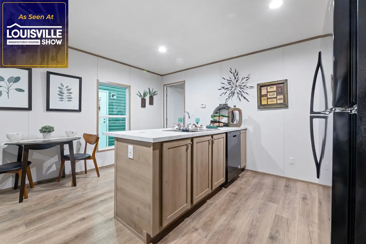 Modern kitchen area with an island, sink, and dishwasher. White walls, light wood cabinetry, and wood-grain flooring. Dining table with chairs and wall decorations are visible. "As Seen At Louisville Manufactured Housing Show" sign in corner.