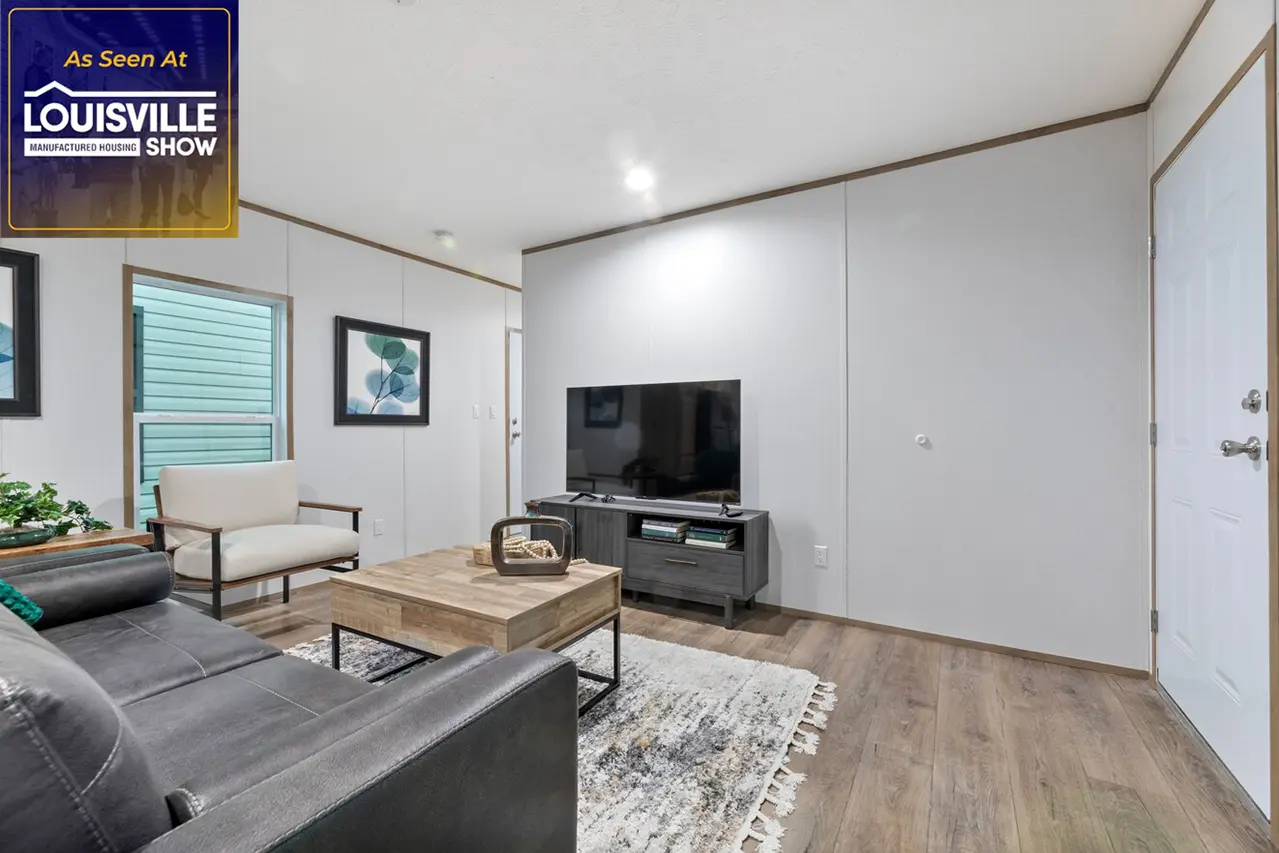 Modern living room with a couch, armchair, coffee table, TV on a stand, wall art, and a rug on wood flooring. A door and interior window are visible. "Louisville Show" banner in corner.