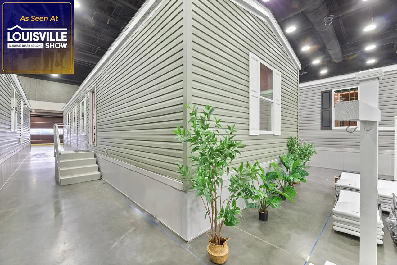 A manufactured home is displayed at the Louisville Manufactured Housing Show. The home is light green with white trim and surrounded by potted plants inside an exhibition hall.