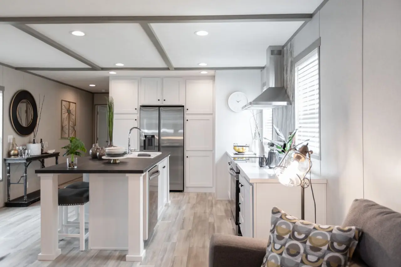 34298bb6-dbfc-40c3-8f3e-3d3cb67ccd28 Modern kitchen with white cabinets, stainless steel appliances, and an island with seating. It features a window with blinds, wood flooring, and a sofa with patterned pillows.