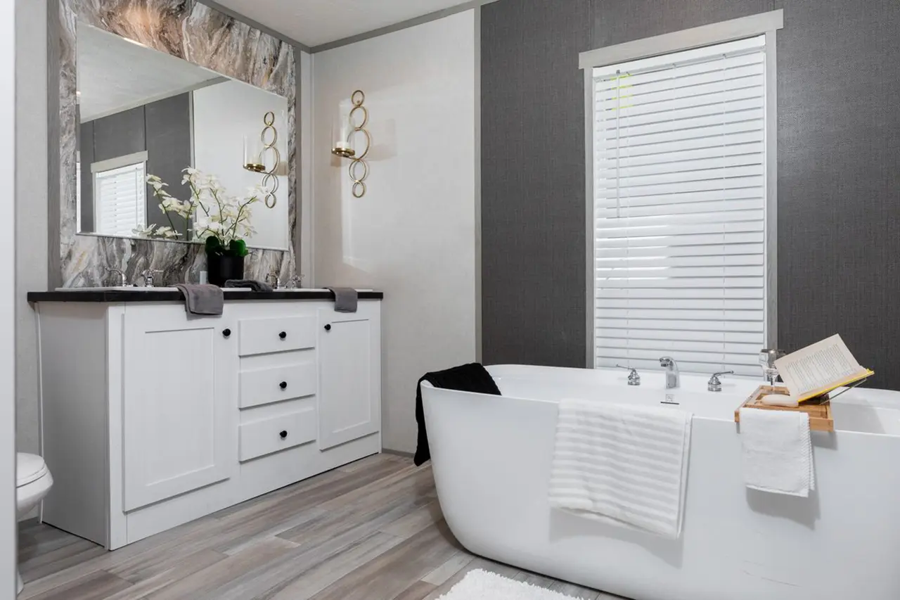 55b7666d-c28e-4232-b08b-cf1779d13cfc Modern bathroom with a freestanding tub, a white vanity with dual sinks, a large mirror, and decorative lighting. A window with blinds is on the gray accent wall.