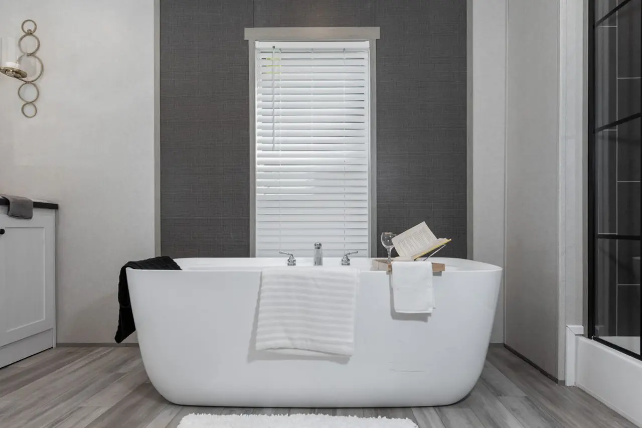 d2fc5b47-2165-4f36-b075-652052240928 Modern bathroom with a freestanding white bathtub, a towel draped over the side, an open book on a bath tray, and a window with closed blinds behind it.