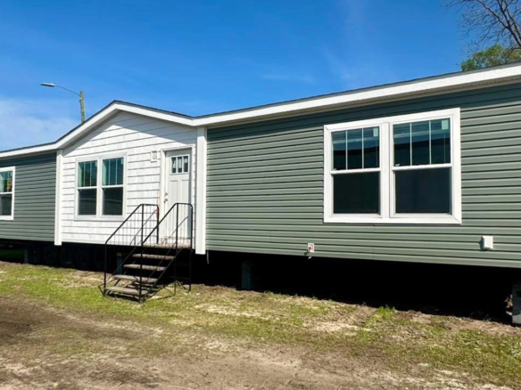A green and white manufactured home with steps leading to the front door, set on a grassy lot under a clear blue sky.