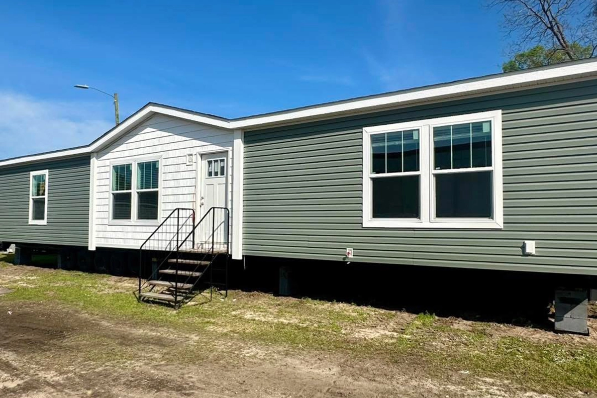 A green and white manufactured home with steps leading to the front door, set on a grassy lot under a clear blue sky.