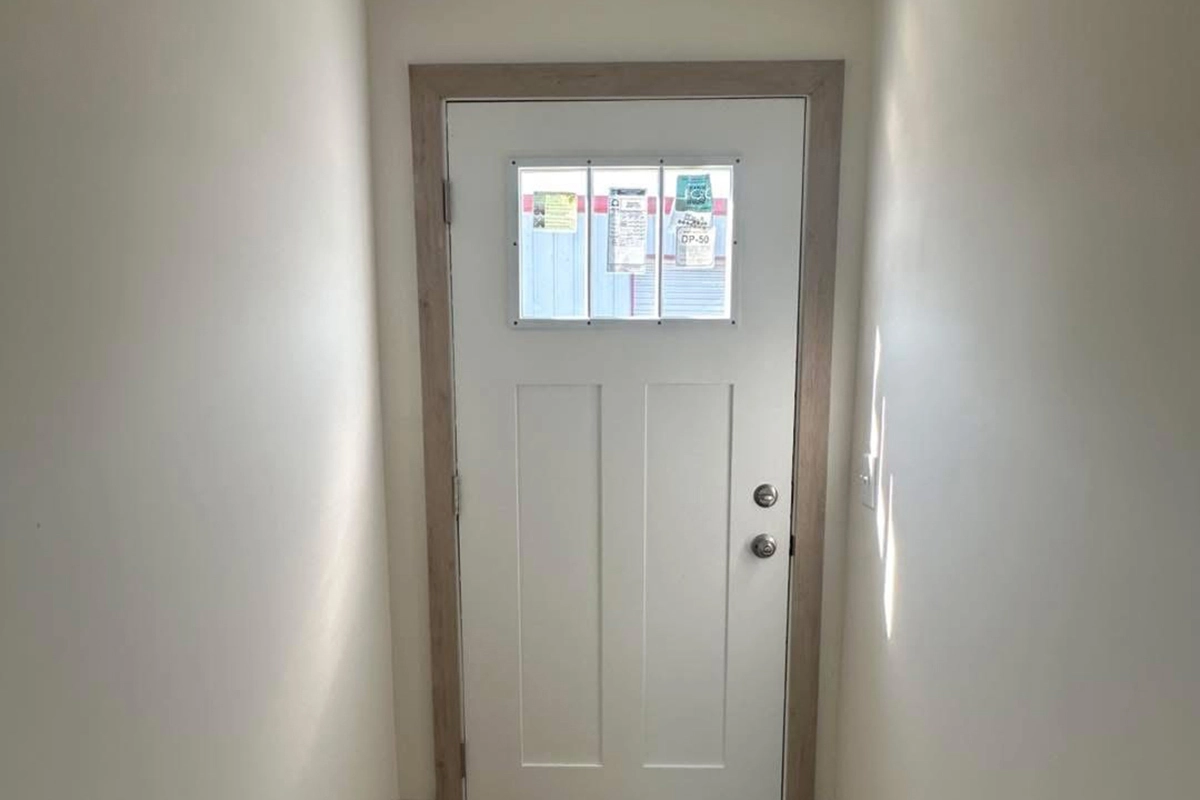 A white door with a wooden frame and a window with four panes, seen from inside a narrow hallway; notices are posted on the window.
