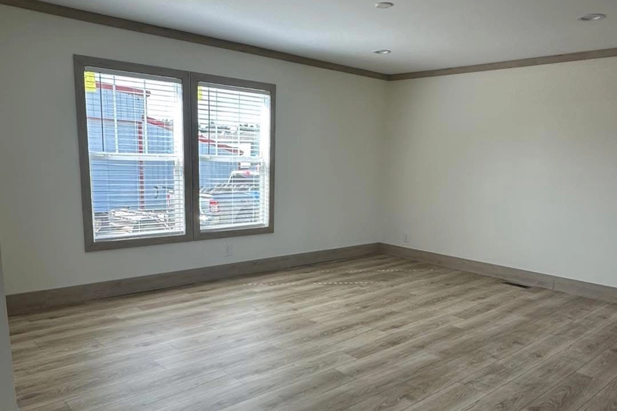 Empty room with light wood flooring, cream-colored walls, two windows with blinds, and natural light coming in. A construction site is visible outside the windows.