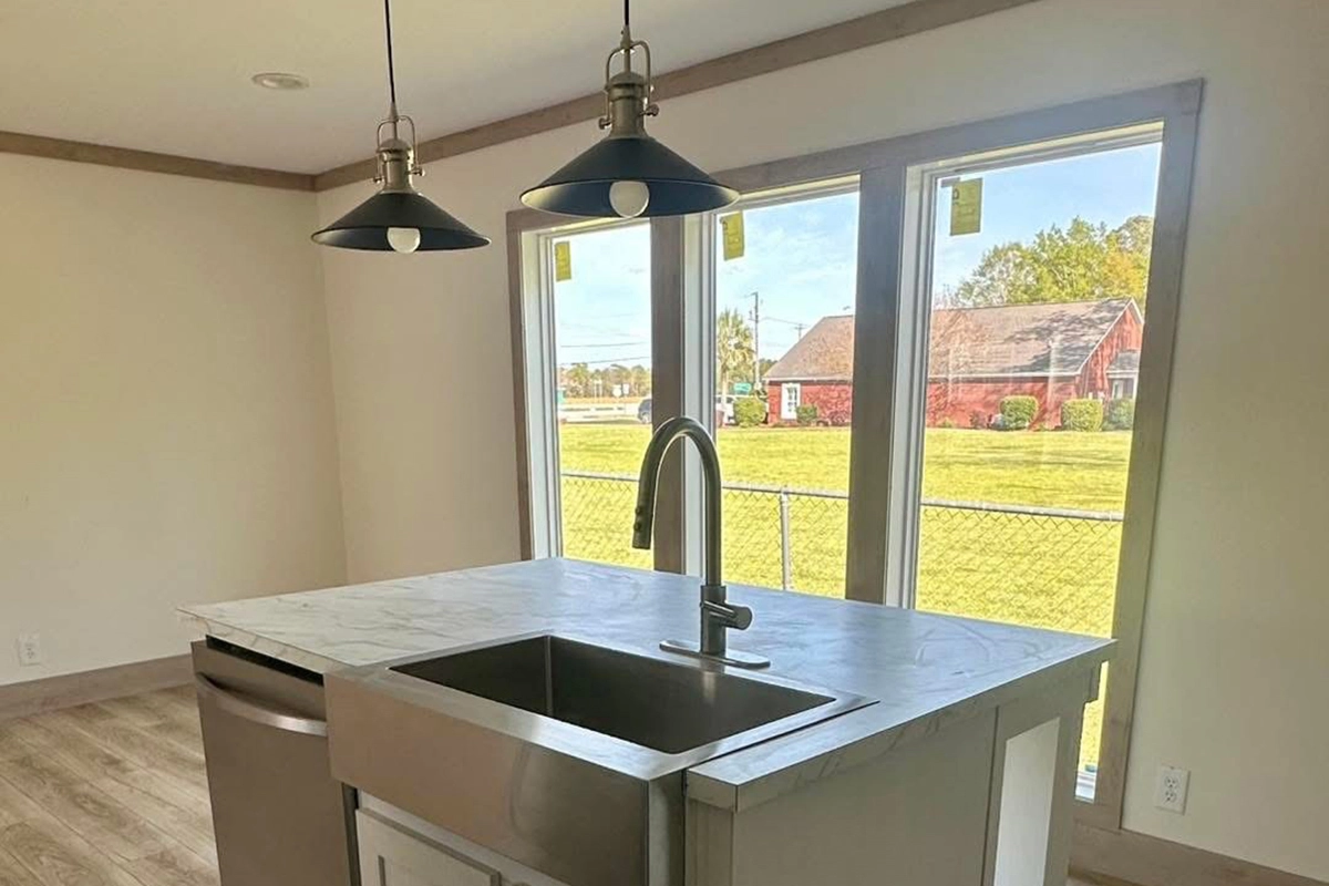 A modern kitchen island with a stainless steel sink and faucet, pendant lights above, and large windows showing a yard and a red barn outside.