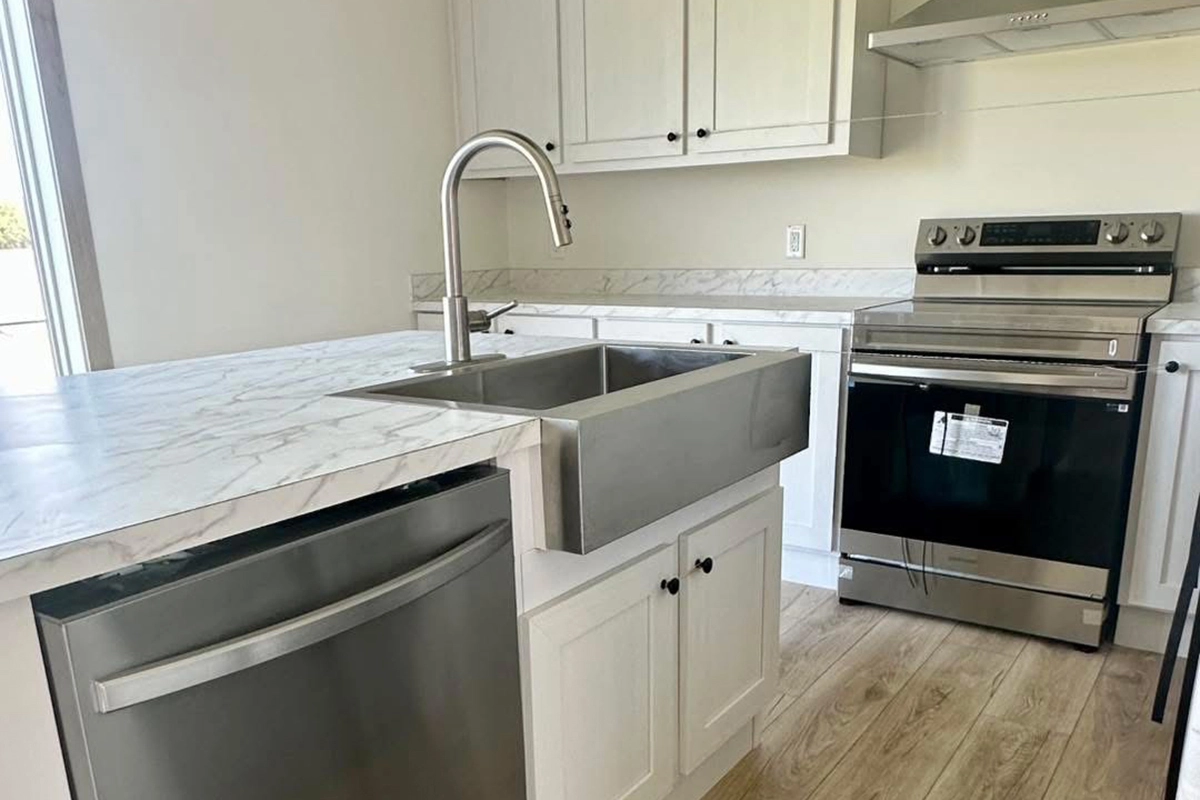 Modern kitchen with white cabinets, stainless steel appliances, a farmhouse sink, marble countertops, and light wood flooring.