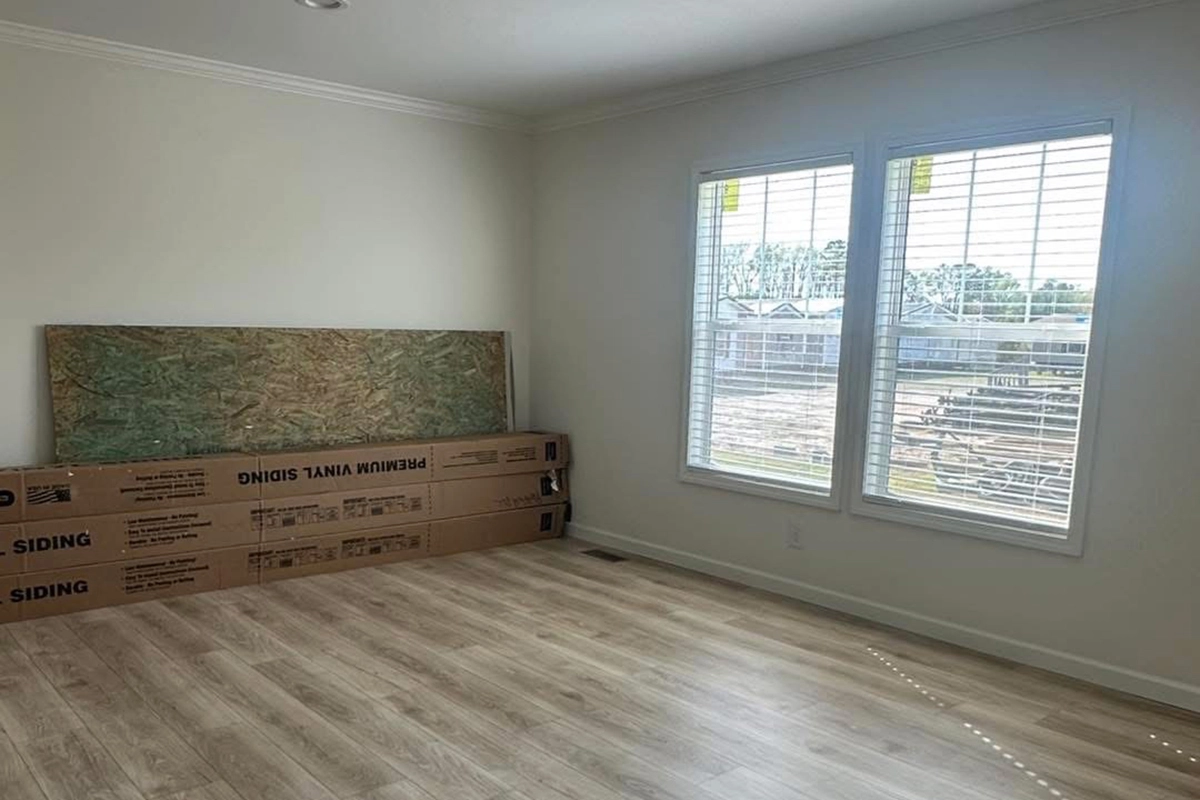 Empty room with light wood flooring, two large windows with blinds, and a boxed item and a covered object leaning against the wall.