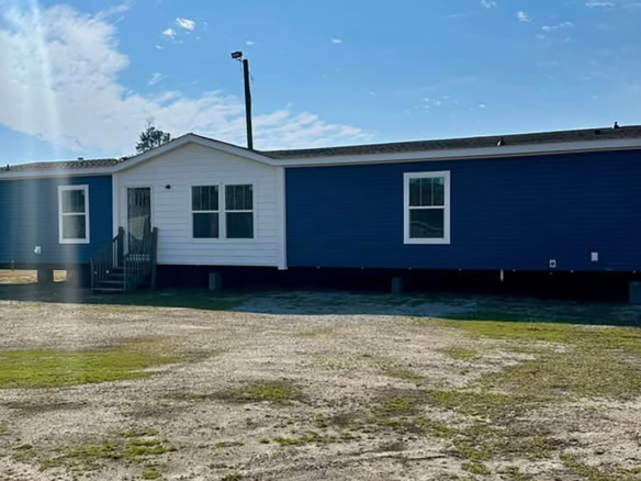 A blue and white manufactured home is elevated on blocks above a patchy dirt and grass yard under a partly cloudy sky.