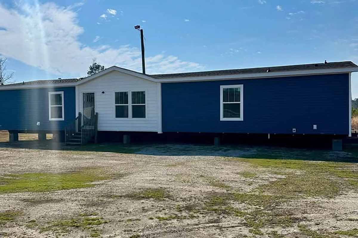 A blue and white manufactured home is elevated on blocks above a patchy dirt and grass yard under a partly cloudy sky.