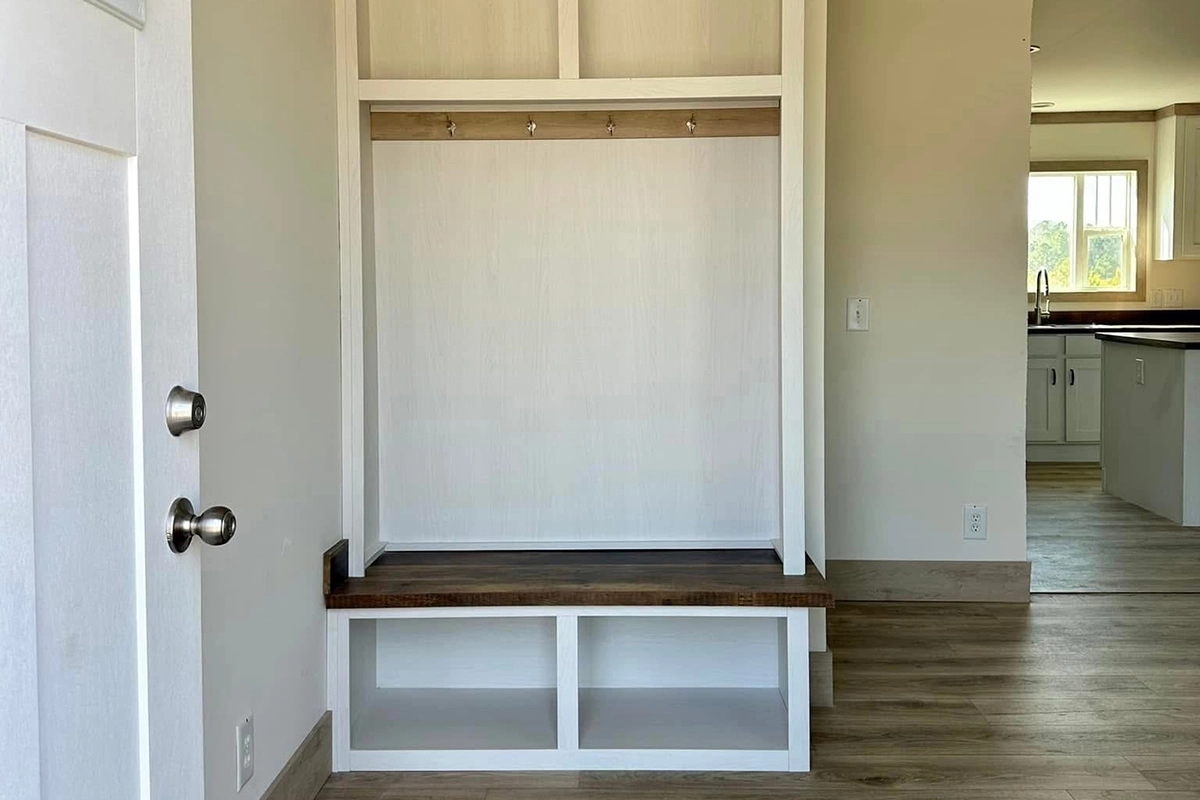 A built-in entryway bench with storage cubbies, hooks, and a wooden seat is situated in a light-colored room with wood flooring near a kitchen.
