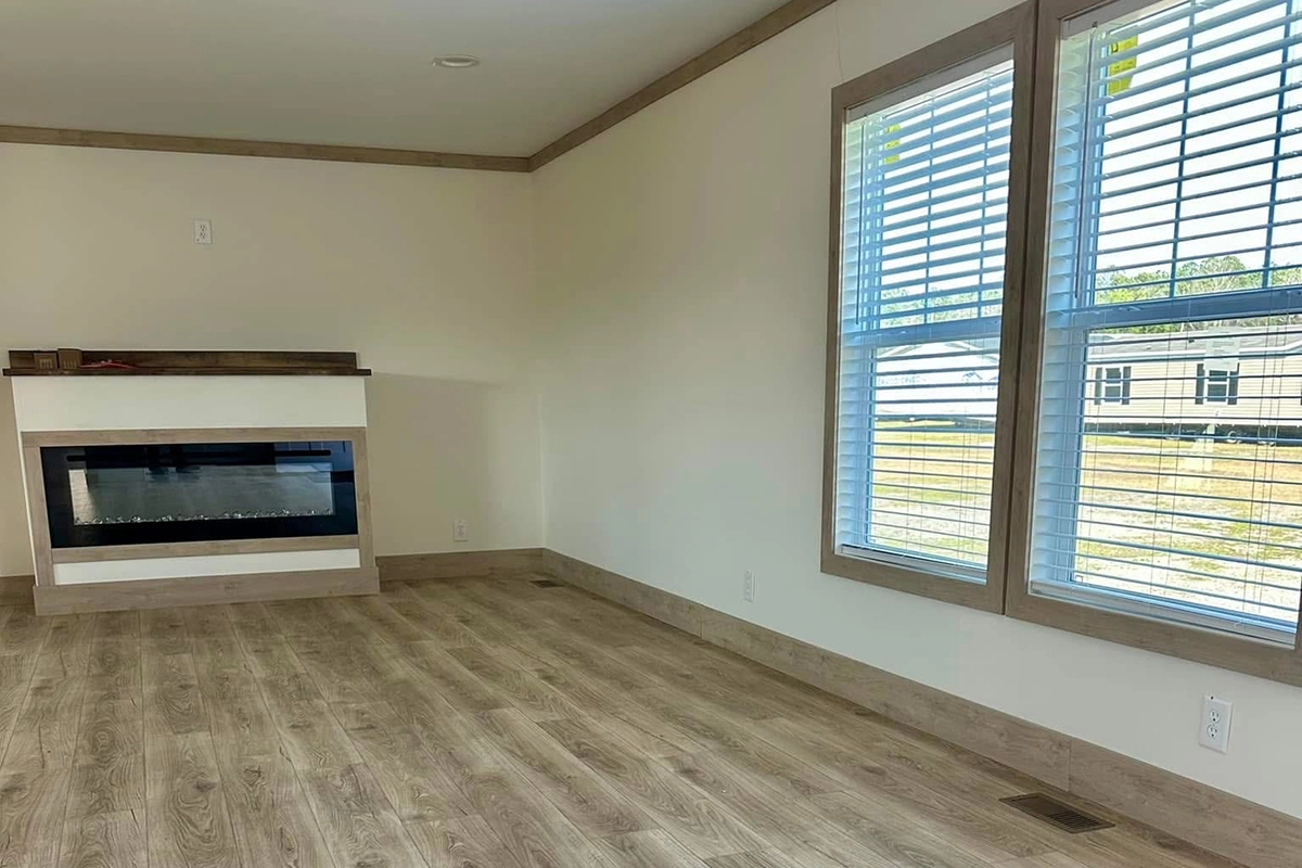 A bright, empty room with light wood flooring, large windows with blinds, and an electric fireplace set into a corner wall.