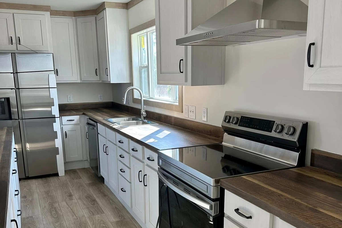 Modern kitchen with white cabinets, stainless steel appliances, electric stove, range hood, double-door refrigerator, and a sink under a window with natural light.