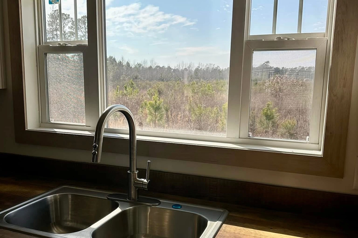 A stainless steel kitchen sink with a faucet is positioned below a large window, overlooking a landscape with trees under a partly cloudy sky.