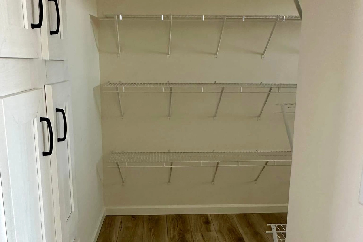 Empty closet with three white wire shelves on the right wall, white cabinets with black handles on the left, and a wooden floor.