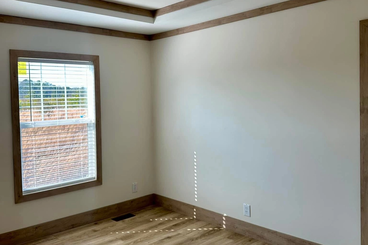 Empty room with light wood trim, beige walls, a large window with blinds, and wood flooring. Sunlight and shadows are visible on the wall and floor.