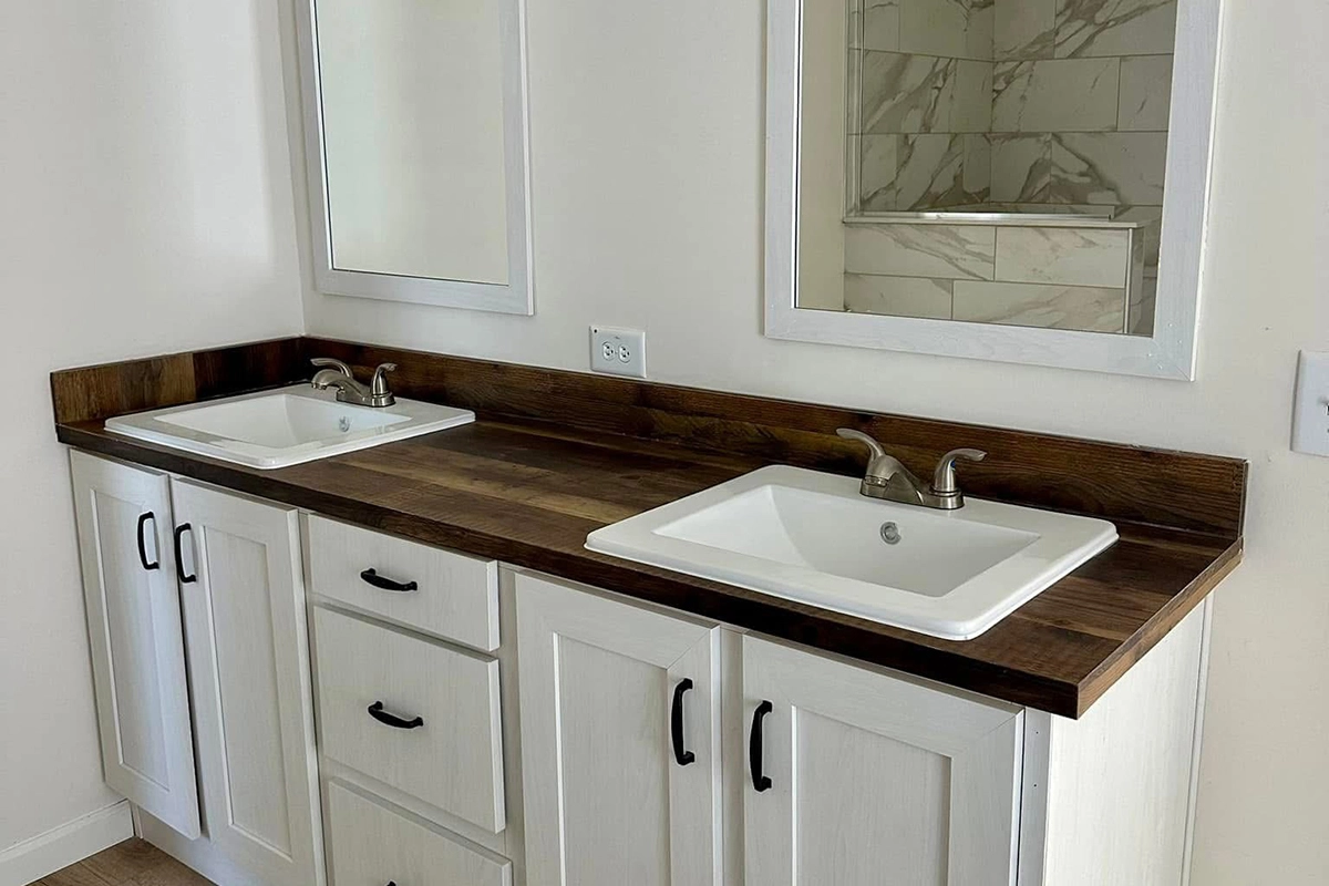 Double bathroom vanity with white cabinets, dark wood countertop, two rectangular sinks with chrome faucets, and mirrors above each sink. Marble tile shower reflected in one mirror.