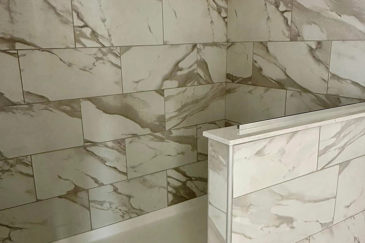 A bathroom wall and half divider covered in large rectangular tiles with a light beige and gray marble pattern.