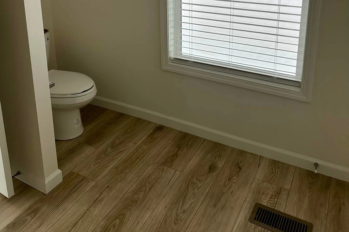 A bathroom with light wood flooring, a white toilet in the corner, a large window with blinds, and a floor vent.