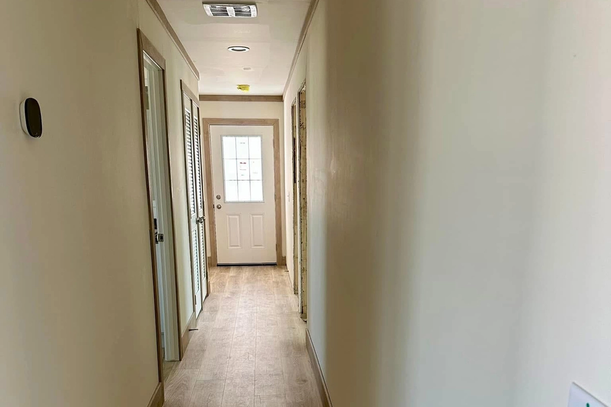 A narrow hallway with light-colored walls and wood trim leads to a closed door with a window at the end; several doors and a thermostat are visible along the hallway.