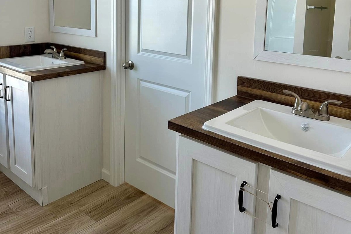 A bathroom with two white vanities, each with a sink, chrome faucets, wood countertops, and a closed white door between them. Light wood flooring.