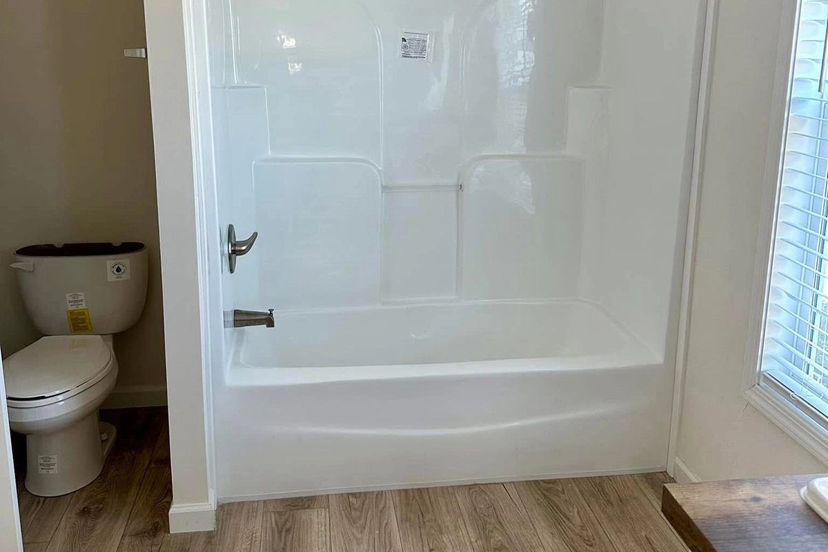 A clean white bathtub and shower combination is next to a toilet in a bathroom with light wood flooring and a window with blinds.