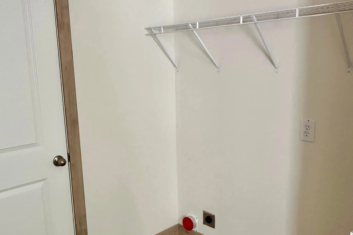 Empty laundry room with a white wall, wire shelf, electrical outlet, plumbing hookups, and a closed door on the left side.