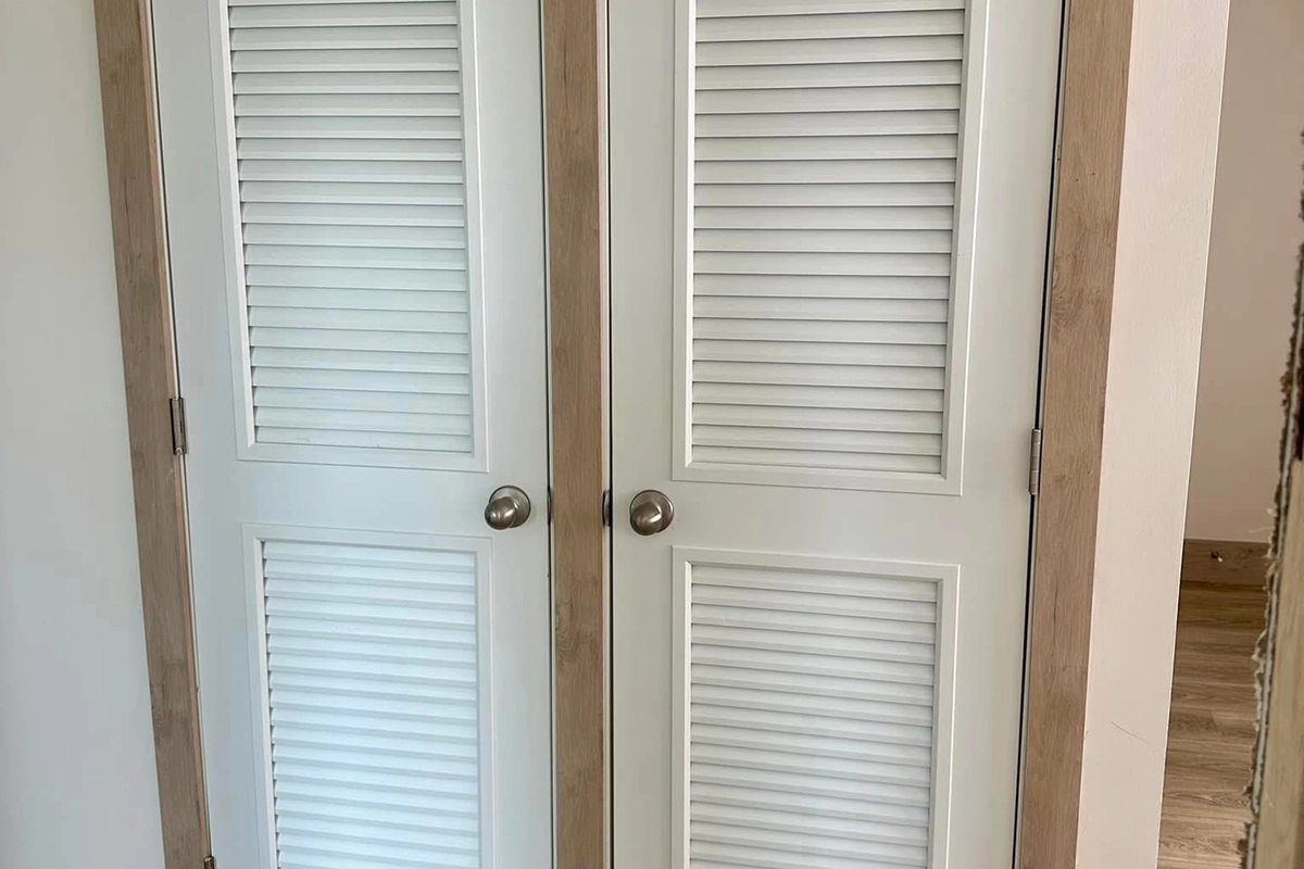 A pair of white louvered closet doors with wooden frames and round metal knobs, set against a light-colored wall and wood floor.