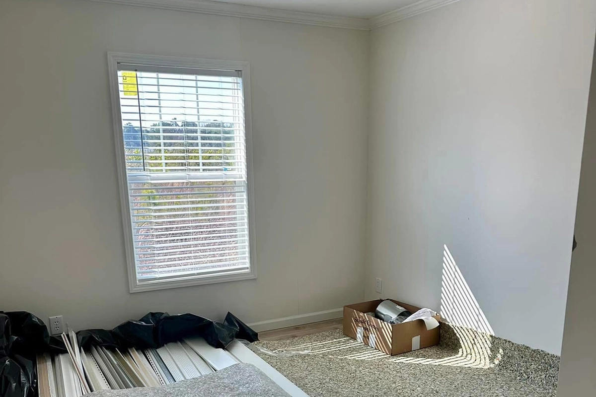 A room with a window, white walls, carpet, some rolled-up materials, black trash bags, and a cardboard box containing various items on the floor. Sunlight casts shadows through the blinds.