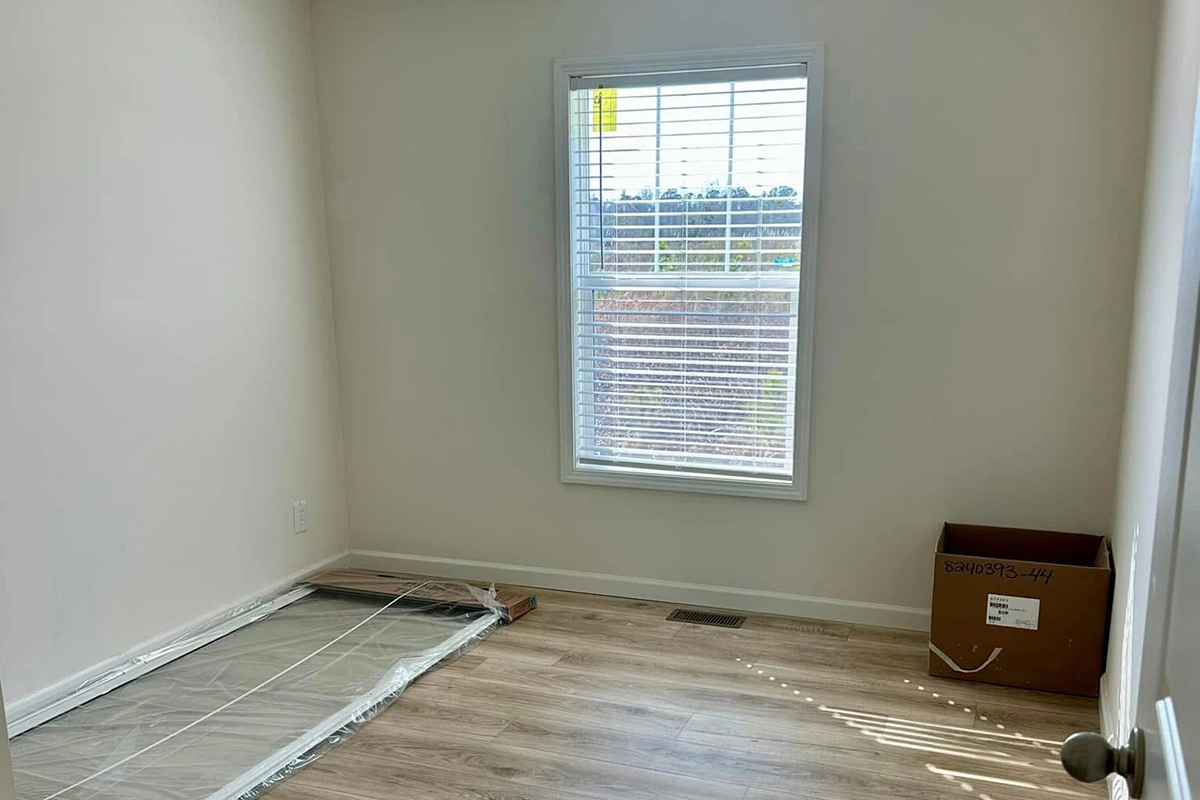 Small empty room with light wood flooring, a window with blinds, a mattress on the floor, and a cardboard box in the corner.