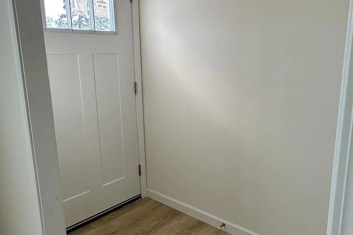 A white entry door with a window at the top is next to a plain, light-colored wall in a small entryway with wood flooring.