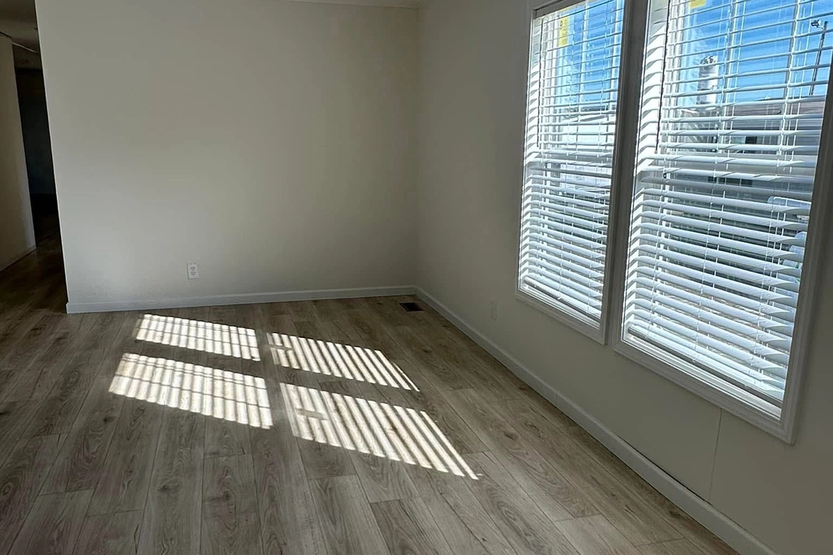 Empty room with light-colored walls and wood flooring, sunlight streaming through two windows with white blinds, casting shadows on the floor.