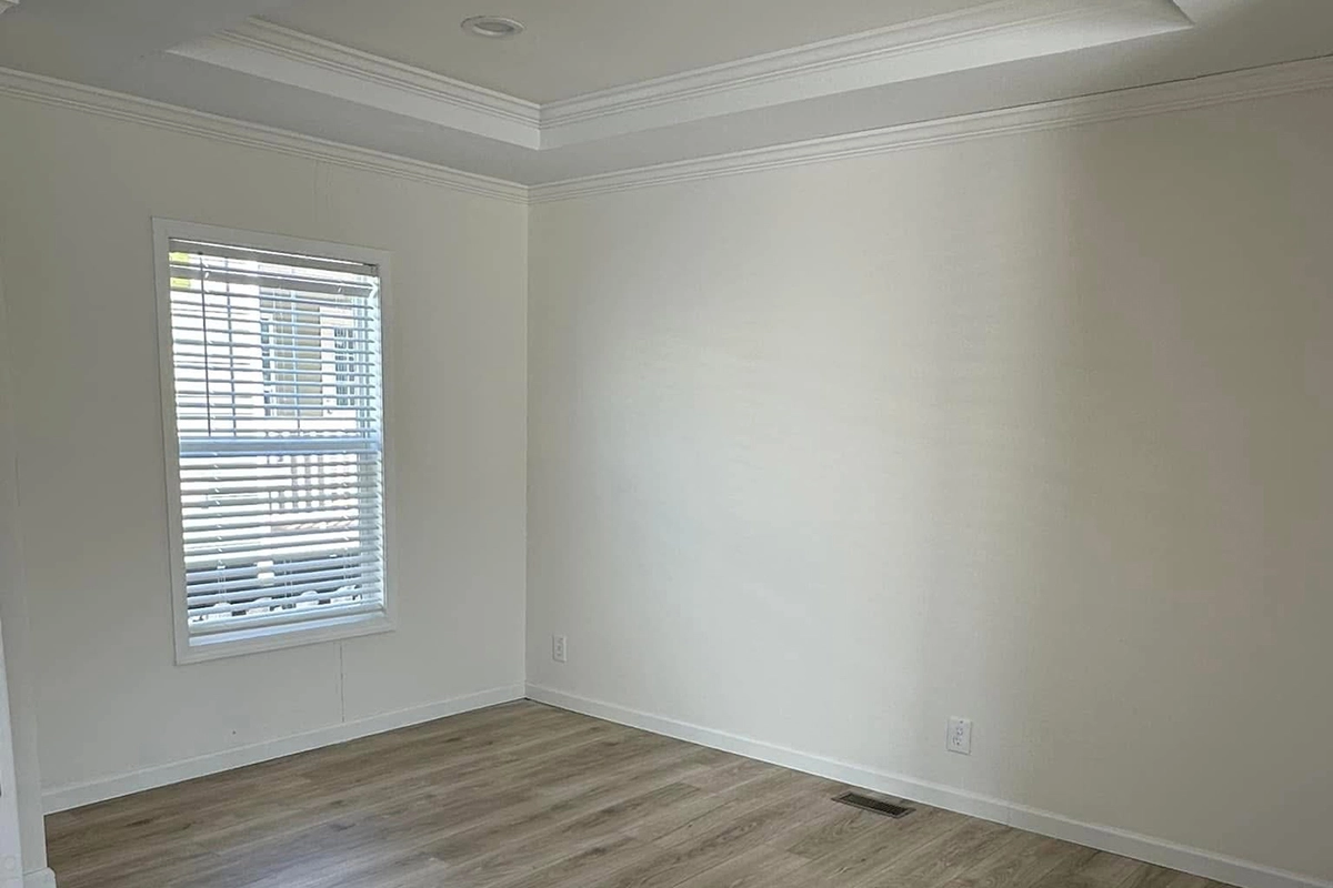 An empty room with light-colored walls, a single window with blinds, wood flooring, and a recessed ceiling.